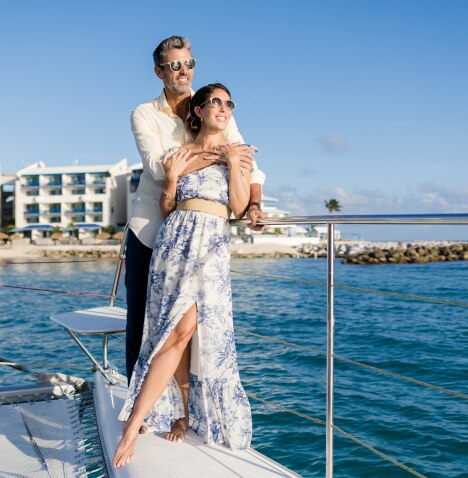 A man and woman in sunglasses, embracing, standing on a boat as it sails across the harbor